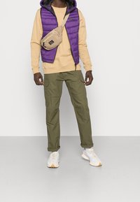 Chaleco acolchado morado sobre una sudadera beige, pantalones cargo oliva y zapatillas claras; con un bolso cruzado de tono neutro.