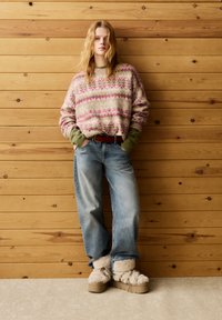 Pletený svetr s růžovými a zelenými vzory, světle modré volné džíny a krémová chlupatá obuv s béžovou platformou, na pozadí dřevěné stěny.
