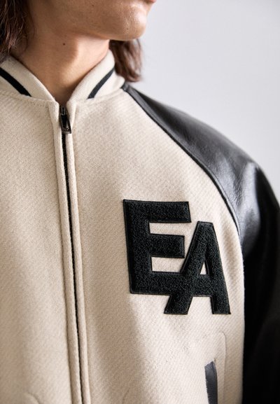 Bežne in črna jakna z zadrgo, teksturirano tkanino in izraženim črnim filcanim logotipom "EA" na sprednji strani.