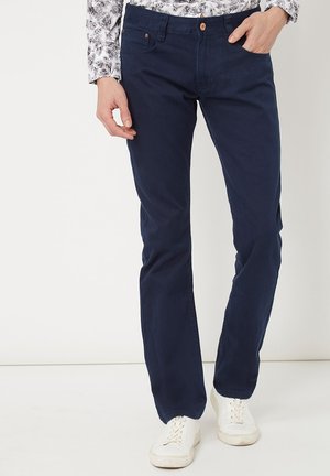 Hombre con pantalones rectos de color azul marino, zapatillas blancas y una camisa de manga larga blanca con patrones de hojas negras, de pie contra un fondo blanco.