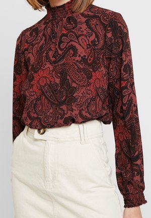 Kvinde iført en vinrød bluse med høj hals og lange ærmer med sort og rødt paisleymønster, stoppet ned i højtaljede off-white bukser.