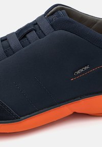 Blue sports shoe with mesh and smooth fabric upper, featuring an orange rubber sole and contrasting stitching. Logo visible on the side.