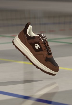 Brauner und schwarzer Sneaker mit weißer Sohle und Schnürsenkeln, der über einem mit farbigen Linien markierten Hallenboden schwebt.
