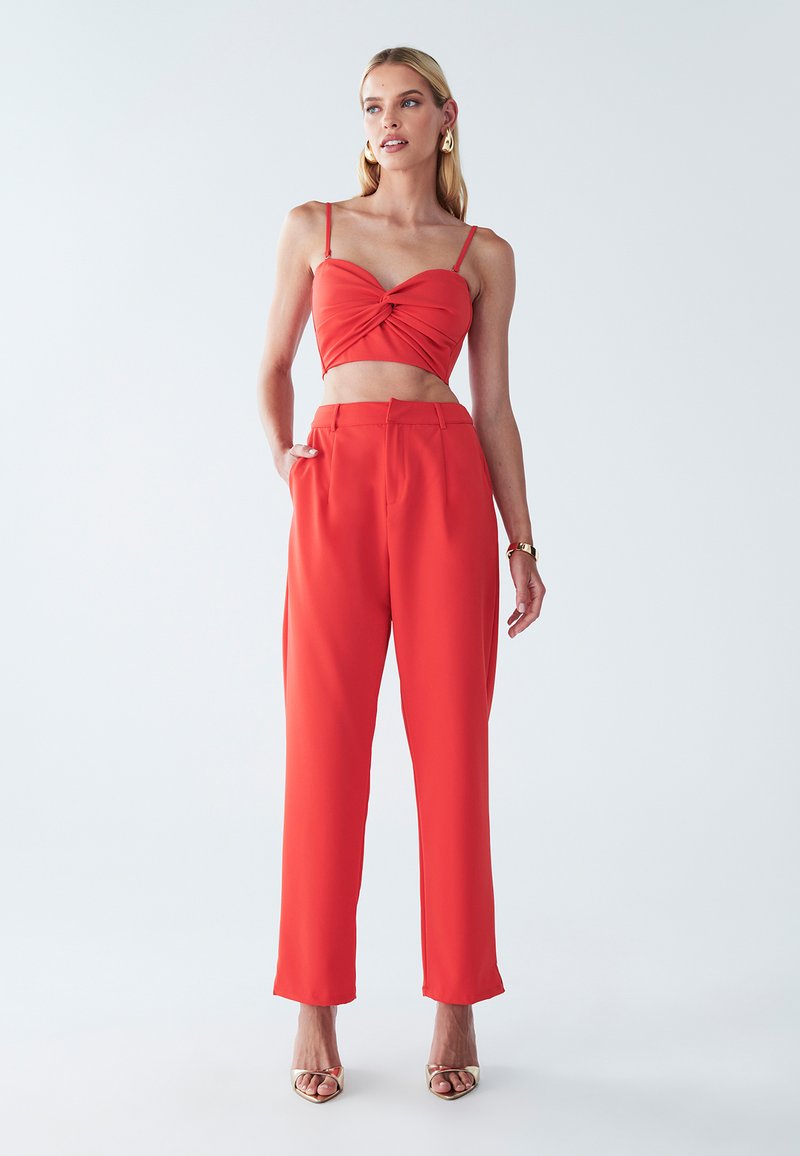 Crveni crop top s čvorom i tankim naramenicama, upareni s crvenim hlačama visokog struka. Glatka, lagana tkanina; moderan siluet.