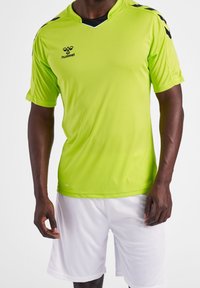 Hombre vistiendo una camiseta deportiva hummel de color verde neón con rayas en forma de chevrón negras en los hombros y pantalones cortos deportivos blancos lisos, de pie contra un fondo blanco.