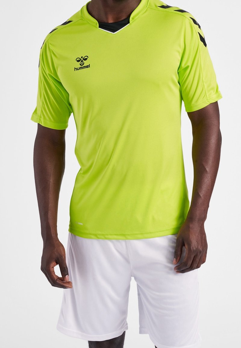 Hombre vistiendo una camiseta deportiva hummel de color verde neón con rayas en forma de chevrón negras en los hombros y pantalones cortos deportivos blancos lisos, de pie contra un fondo blanco.
