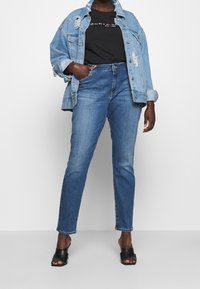 Denimjack met beschadigde details, zwart T-shirt met logo, blauwe jeans en zwarte open teen hakken. Casual outfit.