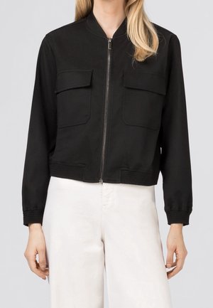 Frau trägt eine schwarze Jacke mit Reißverschluss, zwei Brustklapptaschen und langen Ärmeln, kombiniert mit weißen, hoch taillierten Hosen.