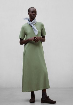 Kvinde iført en lysegrøn maxi-kjole med korte ærmer, gråt tørklæde bundet om halsen, mørke sokker og brune sneakers, stående op ad en ensfarvet væg.