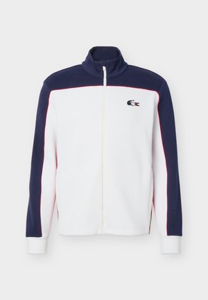 Ζακέτα με πλήρες φερμουάρ σε σκούρο μπλε απόχρωση στο πάνω μέρος και τα μανίκια, λευκό σώμα, λεπτομέρειες με κόκκινες ρίγες και μικρό κεντητό λογότυπο κροκόδειλου στο στήθος.