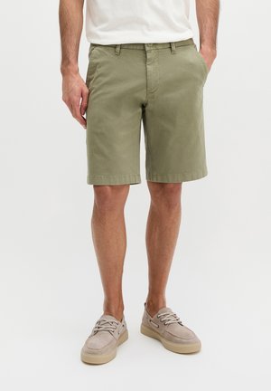 Homme portant un short vert olive jusqu'aux genoux, une chemise blanche et des chaussures décontractées beiges à lacets, debout les mains dans les poches.