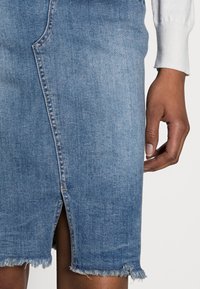 Blue denim skirt featuring a central seam, a frayed hem, and a front slit. Close-up of fabric texture and hand holding the skirt's edge.