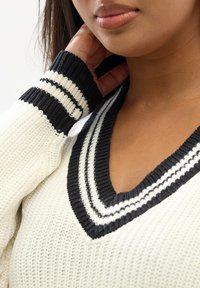 Sweter z dekoltem w serek w kolorze kremowym z czarnymi paskami przy kołnierzu i mankietach; teksturowany dzianinowy materiał; luźny krój, eksponujący detale dekoltu i rękawów.