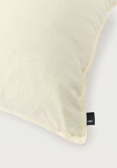 Coussin rectangulaire de couleur crème avec une texture de tissu doux et une petite étiquette noire portant l'inscription "HAY" sur un coin.