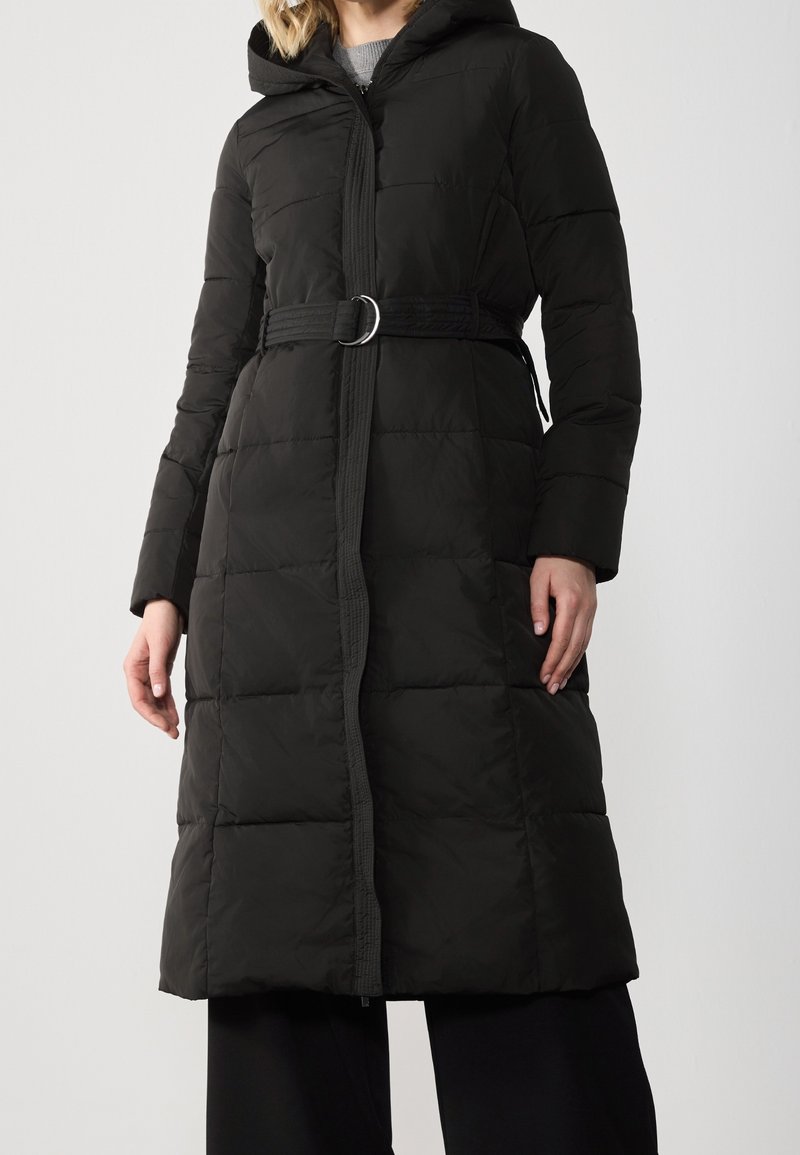 Femme portant un long manteau matelassé noir avec capuche et taille ceinturée, associé à un pantalon noir sur un fond clair uni.