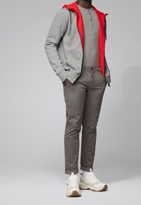 Šedá mikina s kapucí na zip a červeným lemem, světle šedé tričko henley, šedé kalhoty a bílé tenisky s hnědými akcenty. Jednoduchý, neformální outfit.