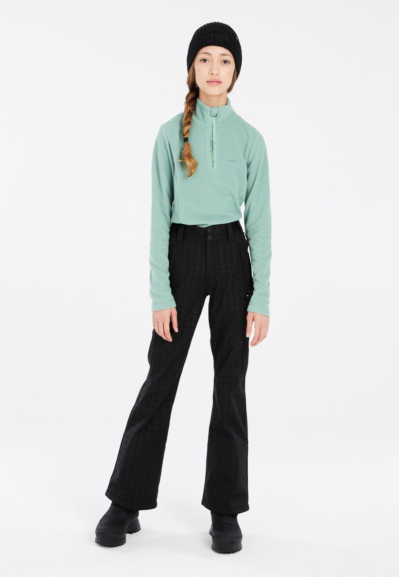 Top in pile verde menta con zip a metà, pantaloni neri a gamba larga con motivo, e un berretto lavorato a maglia nero, abbinati a calzature nere, su uno sfondo bianco.