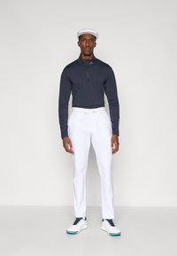 Granatowa koszulka polo z długim rękawem i zamek przy kołnierzyku, białe spodnie oraz białe buty golfowe z granatowymi akcentami. Model nosi białą czapkę.