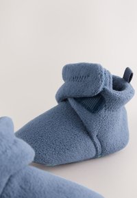 Chaussons en polaire bleue avec un tissu texturé, bout arrondi, bride en Velcro et un accent turquoise sur la semelle. Détail de la doublure intérieure lisse visible.
