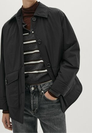 Henkilö pukeutuneena mustaan napilliseen takkiin, jossa alla harmaa- ja valkoraitainen neule ja ruskea poolokaulus, yhdistettynä mustiin farkkuihin.