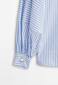 Manche de chemise rayée bleu clair et blanc avec poignet froncé et bouton argenté sur fond blanc.