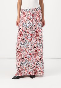 Kvinna i en mönstrad maxikjol med blommor i nyanser av röd, blå och svart, matchad med svarta spetsskor mot en vit bakgrund.