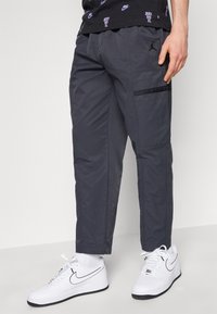 Grijze cargobroek met een getailleerde pasvorm, zijzakken en een gladde textuur, gecombineerd met witte sneakers en een zwart patroonshirt.