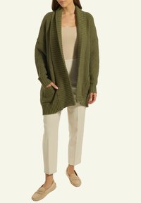 Frau trägt eine olivgrüne, grob gestrickte Strickjacke über einem beigen Top und beigen Hosen mit beigen Mokassins und steht vor einem schlichten hellen Hintergrund.