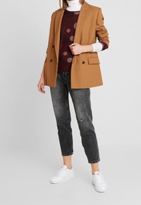 Brązowy blazer z podwójnymi guzikami na wzorzystym swetrze w kolorze burgundowym, zestawiony z szarymi dżinsami z przetarciami i białymi sneakersami.