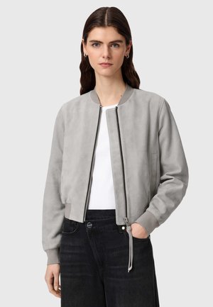 Frau trägt eine hellgraue Bomberjacke über einem weißen Hemd und hoch taillierte schwarze Jeans, steht mit einer Hand in der Tasche vor neutralem Hintergrund.