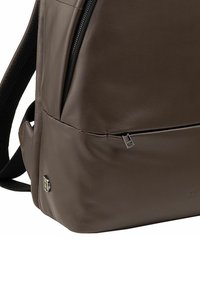 Zaino in pelle marrone con una texture liscia, dotato di una tasca con zip, spallacci regolabili e un dettaglio del logo sul lato.