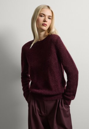 Donna con lunghi capelli biondi che indossa un maglione peloso viola scuro e pantaloni larghi abbinati, in piedi con le mani nelle tasche contro una parete liscia.