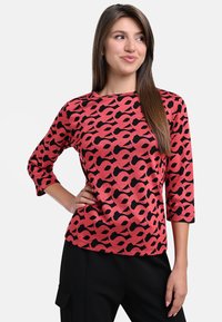 Red and black patterned top made of textured fabric, featuring three-quarter sleeves and a crew neckline. Paired with black trousers.