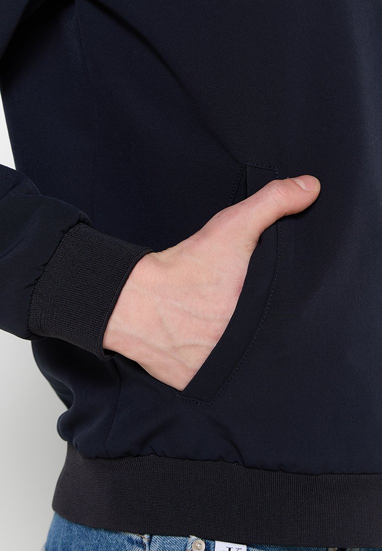 Chaqueta navy con tejido texturizado, cuenta con un bolsillo lateral y puños acanalados. Mano apoyada en el bolsillo, mostrando un diseño ajustado.