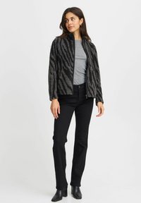 Veste en fausse fourrure noire et grise avec un motif zèbre, fermeture éclair et col montant, associée à une chemise grise et un pantalon noir.