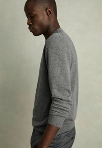 Pull en maille gris avec un col rond, des poignets côtelés et un ourlet ; porté avec un pantalon foncé ; vue de profil sur fond neutre.