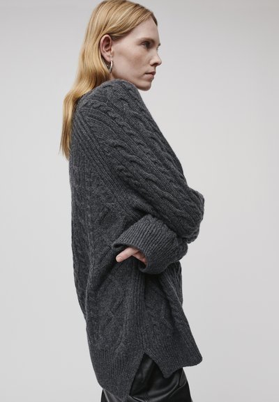 Grauer Kabelstrickpullover mit lockerer Passform, gerippten Bündchen und seitlichen Schlitzen. Weicher Stoff und voluminöses Design; über schwarzen Hosen getragen.
