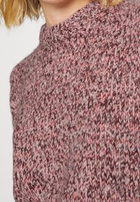 Strickpullover in warmen Rosa- und Grautönen, mit hohem Halsausschnitt und strukturiertem Muster. Der Stoff wirkt flauschig und weich.