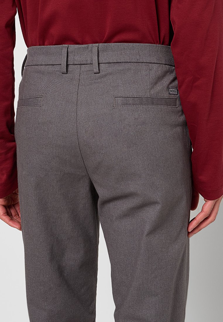 Rückansicht einer Person, die graue, maßgeschneiderte Hosen mit Gürtelschlaufen und Paspeltaschen trägt, kombiniert mit einem langärmligen, dunkelroten Hemd.