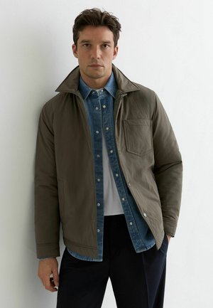 Lekka oliwkowozielona kurtka z wysokim kołnierzem, kieszenią i zatrzaskami, noszona na niebieskiej dżinsowej koszuli i białej koszulce.