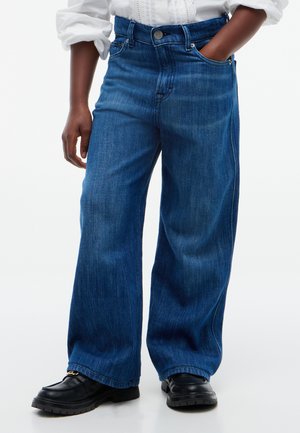 Personne portant un jean bleu ample, des chaussures noires épaisses et une chemise blanche à manches longues avec une main dans la poche.