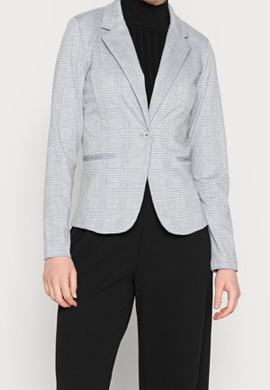 Kvinde iført en tætsiddende grå ternet blazer med enkelt knap over en sort rullekrave og sorte bukser, stående med hænderne afslappet ned langs siderne.