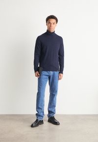 Μπλέ αμάνικο πουλόβερ από μαλακό ύφασμα, φορεμένο με ανοιχτό μπλε τζιν ίσιες γραμμές και μαύρα κορδόνια παπούτσια, κρατώντας ένα μαύρο πορτοφόλι.