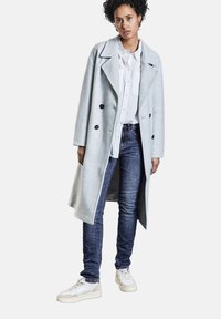 Ljust grå ullkappa med dubbelknäppt design och svarta knappar, kombinerad med en vit skjorta, blå jeans och ljusa sneakers.