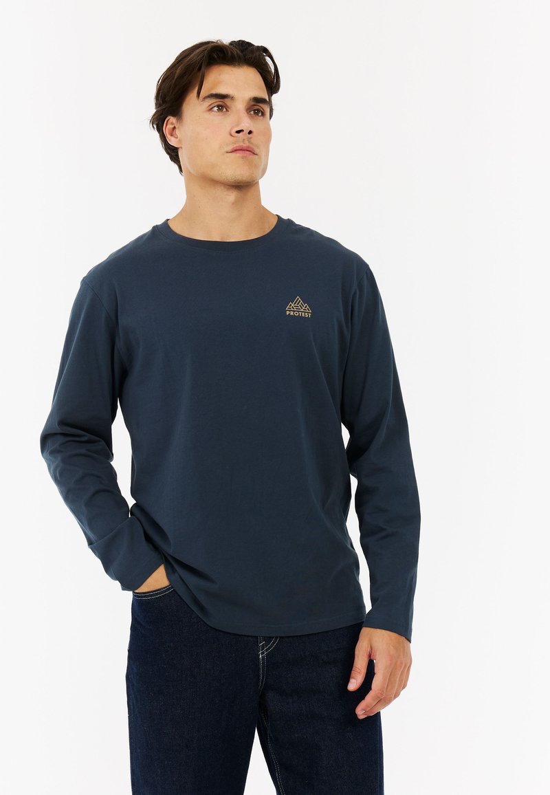 Lange mouwen navyblauwe shirt van katoen, met een ronde hals en een klein gouden berglogo op de linkerborst. Gepaard met donkerdenim jeans.