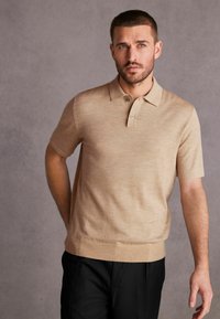 Beige lyhythihainen polo-paita, jossa on hienovarainen rakenne, kolmen napin nappisuljenta ja ribatettu helma. Yhdistetty tummiin housuihin.