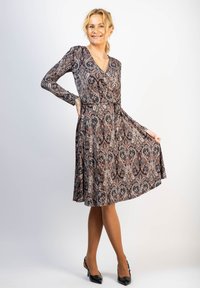 Pitkähihaista, tummasävyistä paisley-kuvioista mekkoa, jossa on kietaisumallinen malli ja levenevä hame. Yhdistetty mustiin kenkiin, vaaleaa taustaa vasten.