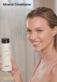 Après-shampoing minéral AHAVA dans une bouteille blanche avec un bouchon marron, de forme cylindrique, et une étiquette avec des accents bleus ; tenu par une personne souriante.
