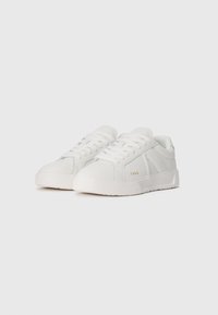 Et par hvide low-top sneakers med teksturerede såler, snørebånd og diskret branding på siden, mod en ensfarvet baggrund.