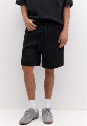 Persoană purtând pantaloni scurți negri cu șnur, șosete albe, adidași gri, tricou negru, inele și o brățară, stând pe un fundal alb.